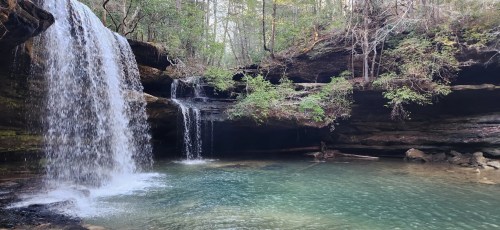 A serene waterfall cascades into a clear pool, surrounded by lush greenery and rocky cliffs.