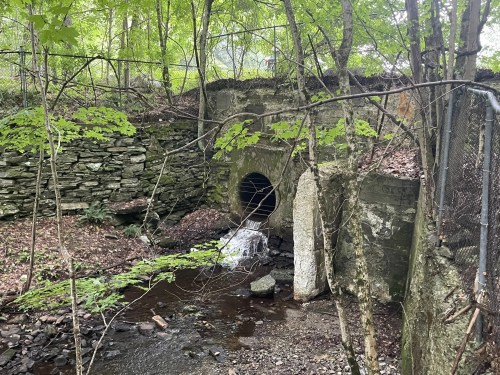 A small stream flows from a stone tunnel, surrounded by trees and rocky terrain. Fencing is visible in the background.