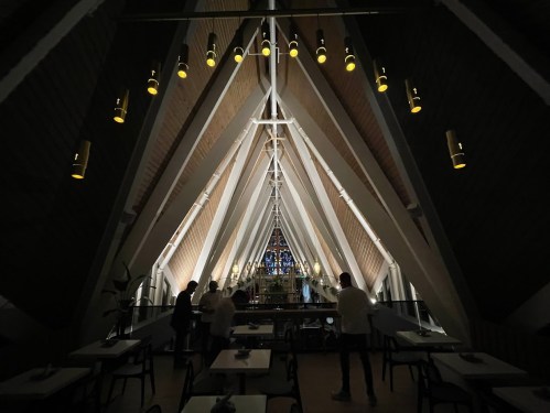 Interior of a modern building with a high, triangular ceiling and warm lighting, featuring people and tables below.
