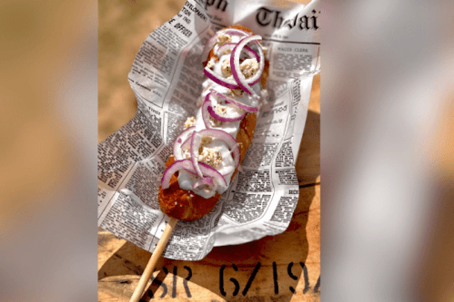 A close-up of a corn dog topped with onions and sauce, served on newspaper.