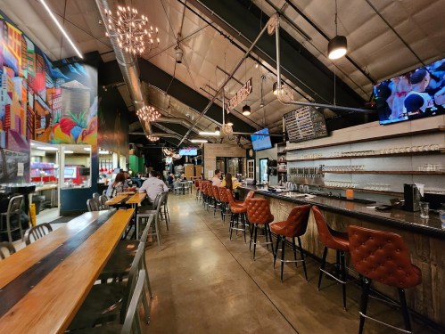 A modern bar interior with high ceilings, colorful murals, and seating at the counter and tables, featuring TVs and pendant lights.