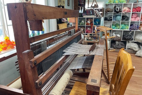 A wooden loom in a craft studio, with woven fabric and colorful yarns displayed in the background.