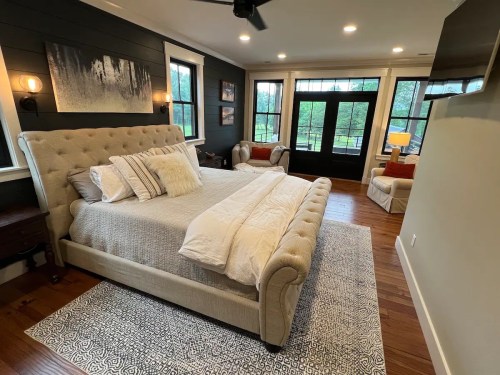 Cozy bedroom featuring a tufted bed, armchairs, and large windows overlooking a green view. Warm, inviting decor.