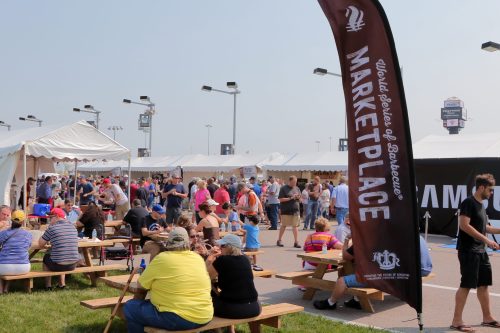 Crowd enjoying a barbecue festival with tents and a large banner reading "Marketplace" in a sunny outdoor setting.