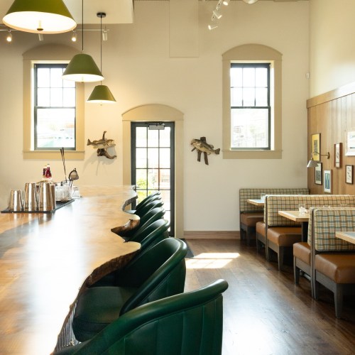 A cozy restaurant interior featuring a wooden bar, green chairs, and booths with plaid upholstery. Natural light from windows.