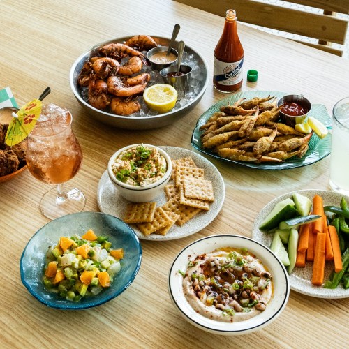 A vibrant spread of seafood, dips, fresh vegetables, and drinks on a wooden table, showcasing a variety of colorful dishes.