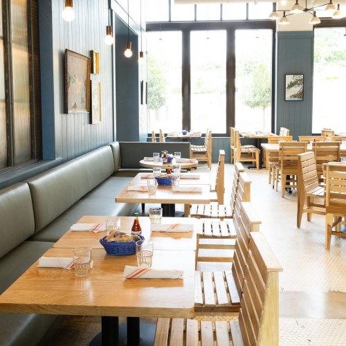 Bright, modern restaurant interior with wooden tables, benches, and large windows letting in natural light.