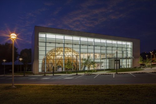 Modern building with large glass windows and a wooden dome structure, illuminated at dusk with a cloudy sky.