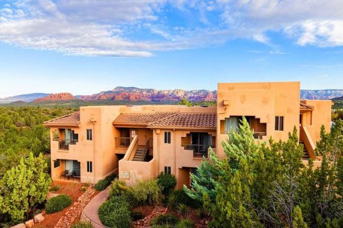 A desert-style hotel nestled among trees, with red rock formations and blue skies in the background.