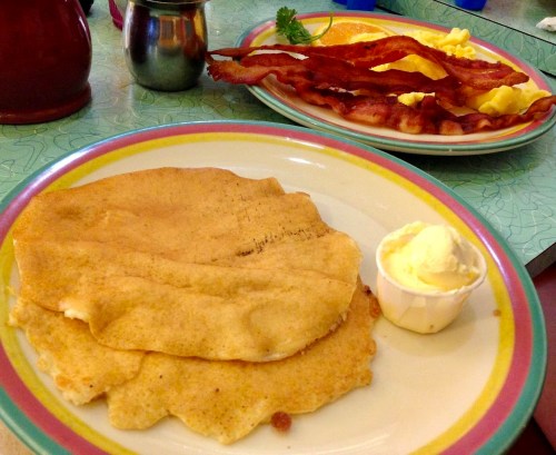 A plate with two flatbreads and a small cup of butter, alongside a plate of bacon and scrambled eggs in the background.