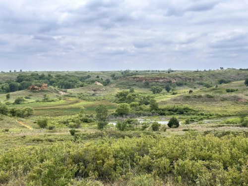 A scenic landscape featuring rolling hills, green vegetation, and a cloudy sky, with a small body of water in the foreground.