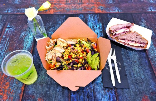 A takeout meal featuring a salad with grilled chicken, corn, and avocado, alongside a sandwich and a drink.