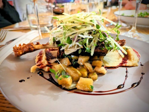A gourmet dish featuring a lamb chop atop apple compote, garnished with fresh greens and drizzled with sauce.