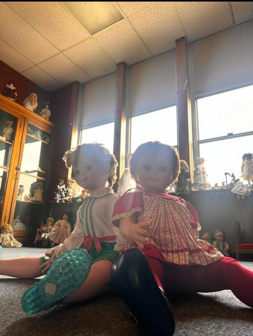 Two vintage dolls sit on a carpeted floor, with sunlight streaming through windows in a room filled with other dolls.