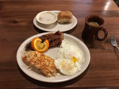 A breakfast plate with eggs, bacon, hash browns, an orange slice, a biscuit, and a cup of coffee on a wooden table.