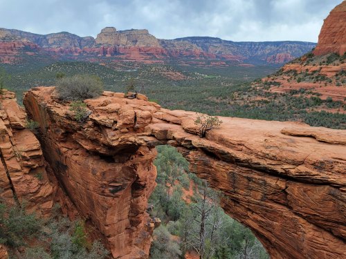 A rocky arch spans a canyon, surrounded by red cliffs and green trees under a cloudy sky.