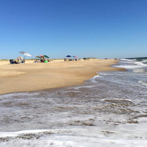 A sunny beach scene with umbrellas, people relaxing, and gentle waves lapping at the shore under a clear blue sky.
