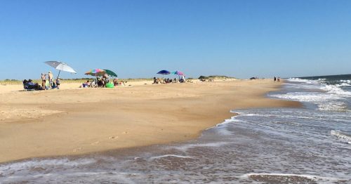 A sunny beach scene with umbrellas, people relaxing, and gentle waves lapping at the shore under a clear blue sky.