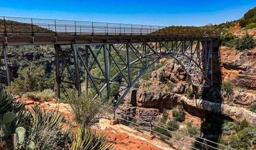 A metal arch bridge spans a canyon, surrounded by rocky cliffs and greenery under a clear blue sky.