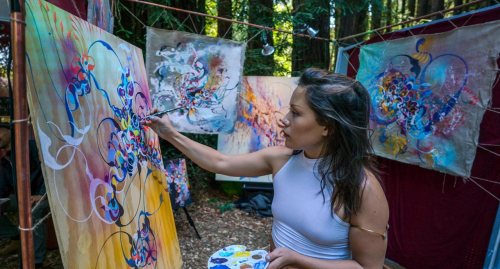 A woman paints on a canvas in a forest, surrounded by colorful abstract artworks hanging nearby.
