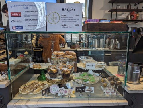A bakery display case filled with various pastries, cookies, and gluten-free options, with prices listed above.