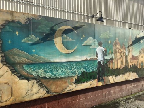 A vibrant mural depicting a man painting a scenic landscape with mountains, ocean, and a crescent moon.