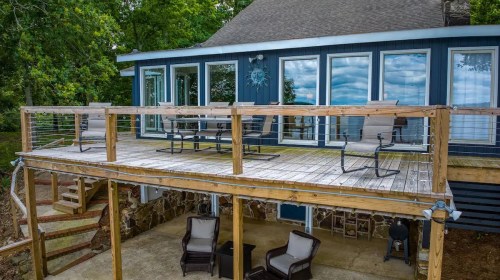A wooden deck with seating overlooks a lake, attached to a blue house with large windows and a stone foundation.