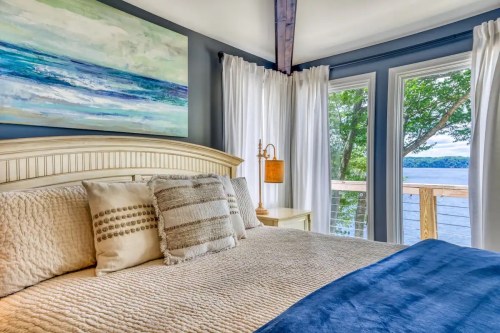 Cozy bedroom with a large bed, decorative pillows, and a view of a lake through large windows.