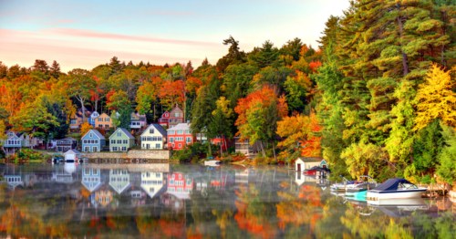 Colorful houses reflect in a calm lake surrounded by autumn trees, creating a serene fall landscape.