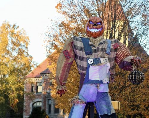 A large scarecrow with a pumpkin head stands in front of a historic building, surrounded by autumn trees.