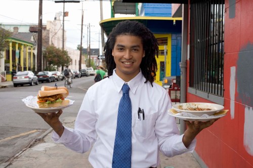 A smiling young man in a white shirt and tie holds two plates of food outside a colorful restaurant.