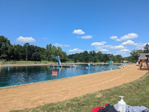 A sunny day at a swimming area with a sandy beach, diving board, and clear blue water under a bright blue sky.