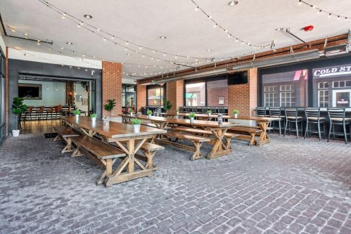 A spacious restaurant interior with wooden tables, string lights, and a brick floor, featuring a bar area in the background.