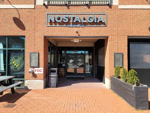 Entrance of Nostalgia Brewing Co. featuring a brick facade, large sign, and outdoor seating area.