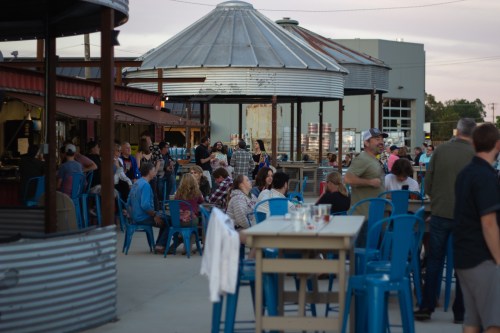 A lively outdoor gathering with people seated at blue chairs, enjoying food and drinks under a metal-roofed structure.