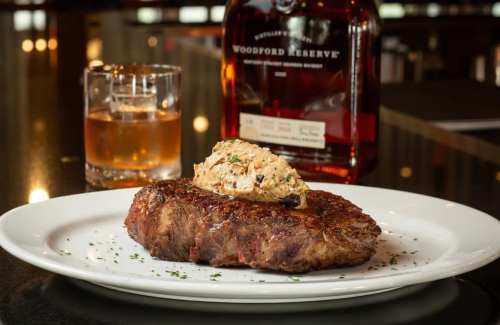 A juicy steak topped with creamy sauce, served with a glass of whiskey and a bottle of Woodford Reserve in the background.