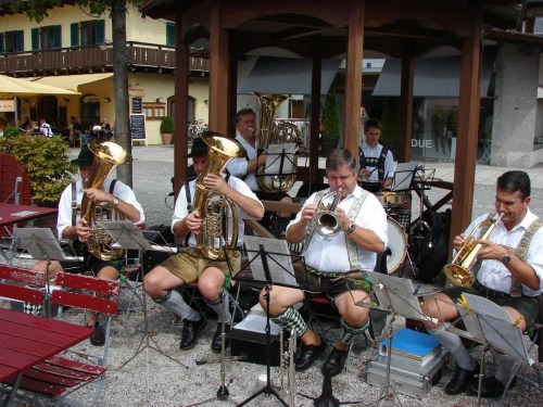 A brass band performs outdoors, dressed in traditional attire, with instruments and sheet music in a lively setting.