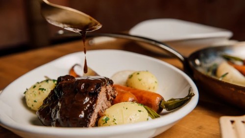 A plate of meat with rich sauce, surrounded by potatoes and carrots, garnished with herbs.