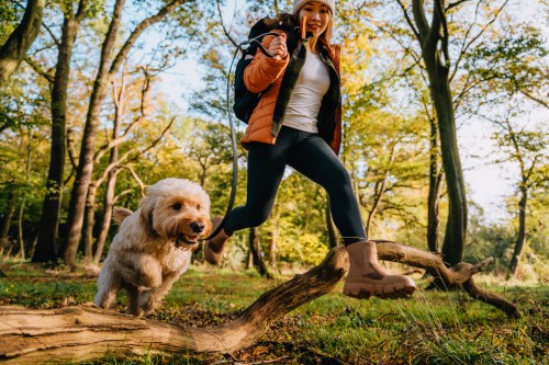 A woman in outdoor gear joyfully runs with a dog through a sunlit forest, surrounded by trees and autumn foliage.