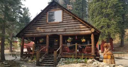 A rustic log cabin lodge surrounded by trees, featuring a porch with rocking chairs and a wooden bear sculpture.