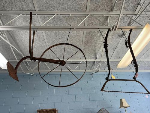Two vintage farming tools hanging from a ceiling, including a plow and a wheel, against a blue wall.