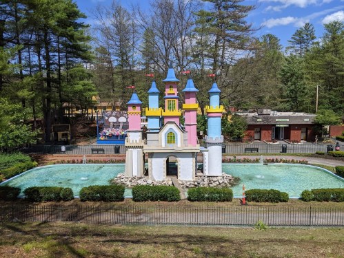 Colorful castle structure surrounded by water, with trees and a playground in the background under a blue sky.