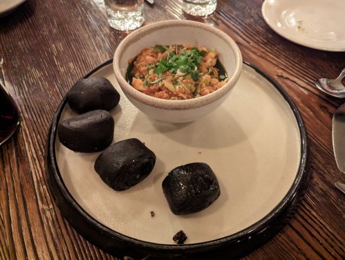 A bowl of savory dish topped with herbs, accompanied by three black rolls on a round plate. Wooden table setting.