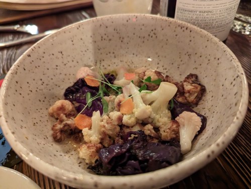 A speckled bowl filled with roasted cauliflower, red cabbage, and garnished with herbs and small vegetables.