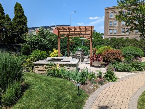 A lush garden featuring a wooden pergola, stone seating area, and vibrant flowers under a clear blue sky.