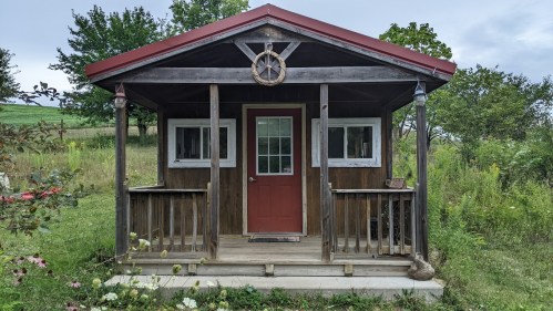 A small wooden cabin with a red roof, front porch, and a decorative wheel above the door, surrounded by greenery.