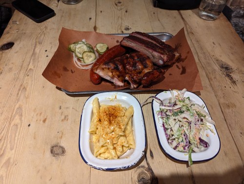 A wooden table with a platter of smoked meats, pickles, and two sides: macaroni and cheese, and coleslaw.