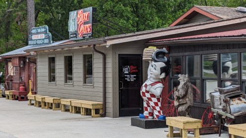 A retro diner exterior featuring a large mascot statue, wooden benches, and a sign for "Kitty's Cafe."