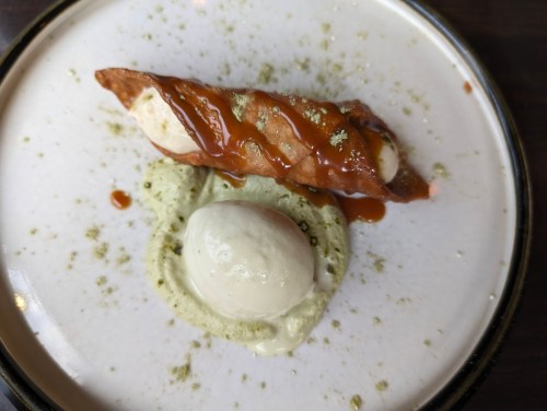 A dessert plate featuring a cannoli filled with cream, drizzled with caramel, and a scoop of green tea ice cream.