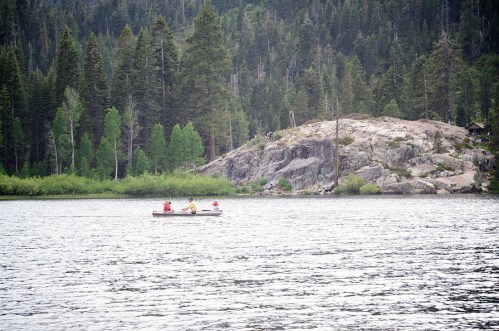 A small boat with people paddling on a calm lake, surrounded by lush green trees and rocky terrain.